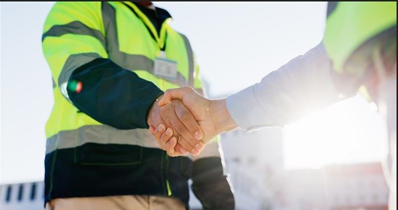Das Bild zeigt einen Mann mit Sicherheitsweste, der einem Mann mit Hemd die Hand gibt.
